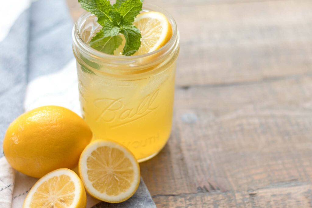 closeup of a glass wide mouth mason jar with iced homemade lemonade garnished with a fresh mint sprig and lemon slice. A whole and halved lemon sitting on a blue and white flour sack towel next to the cup of lemonade