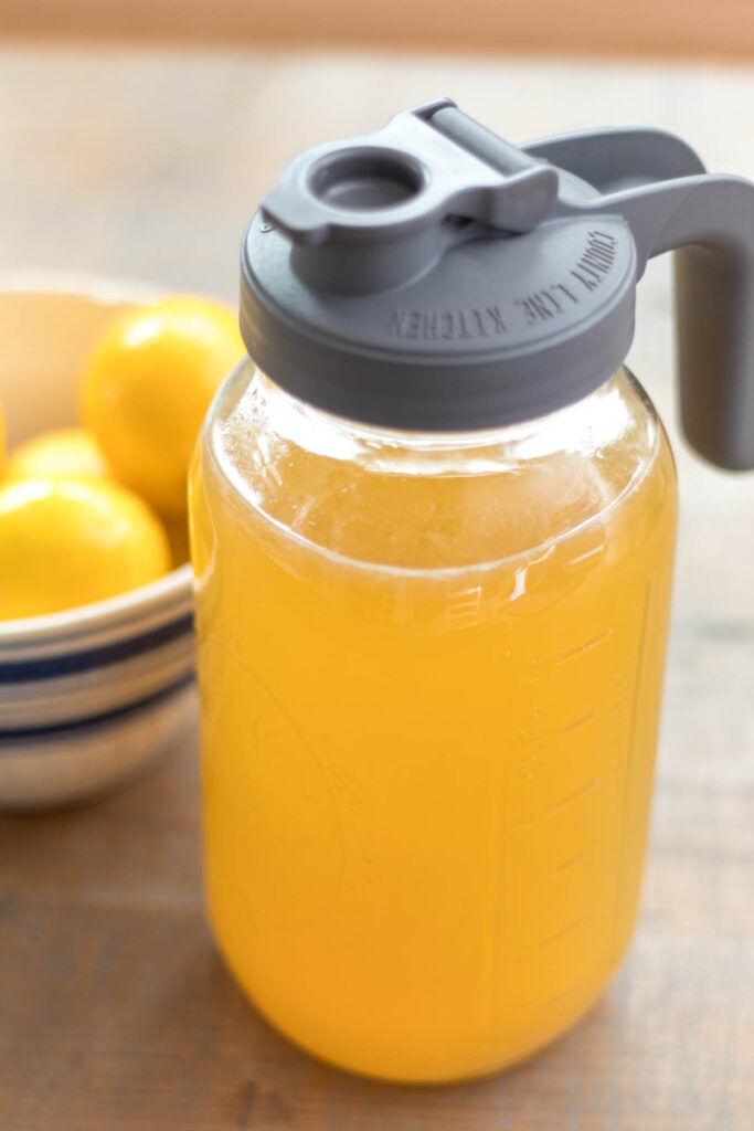 half gallon mason jar with a pitcher lid handle full of homemade honey lemonade and a white with blue stripe antique stoneware bowl filled with lemons in the background