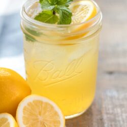closeup of a glass wide mouth mason jar with iced homemade honey lemonade garnished with a fresh mint sprig and lemon slice. A whole and halved lemon sitting on a blue and white flour sack towel next to the cup of lemonade