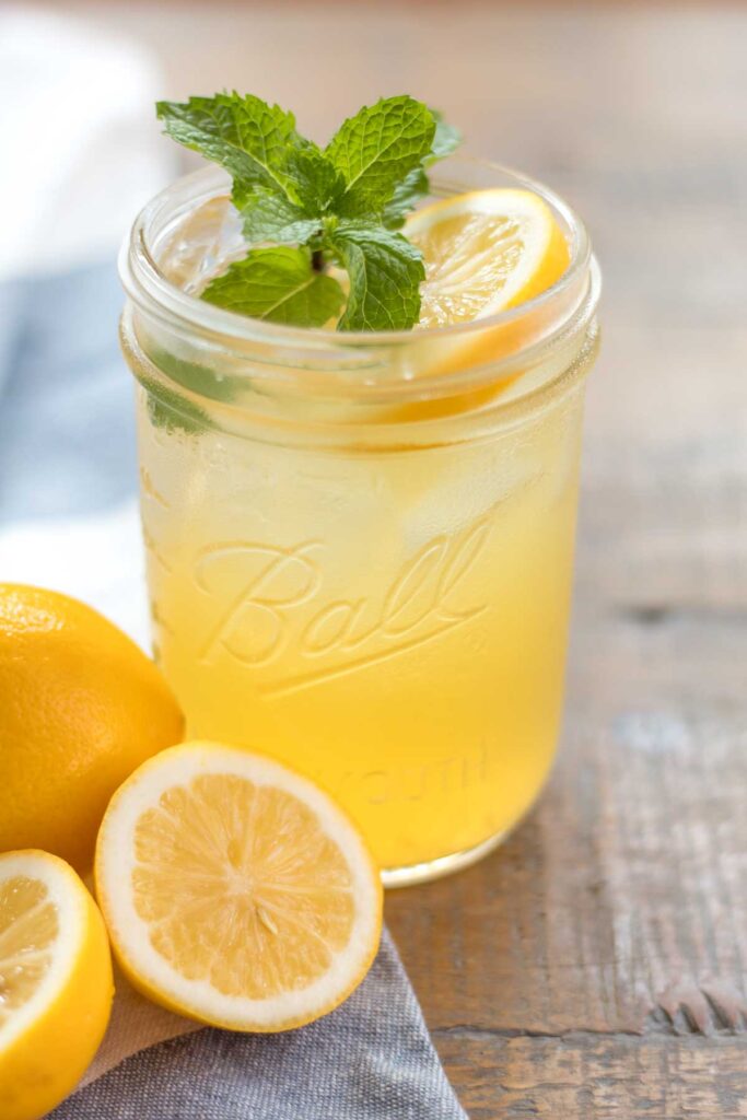 closeup of a glass wide mouth mason jar with iced homemade honey lemonade garnished with a fresh mint sprig and lemon slice. A whole and halved lemon sitting on a blue and white flour sack towel next to the cup of lemonade