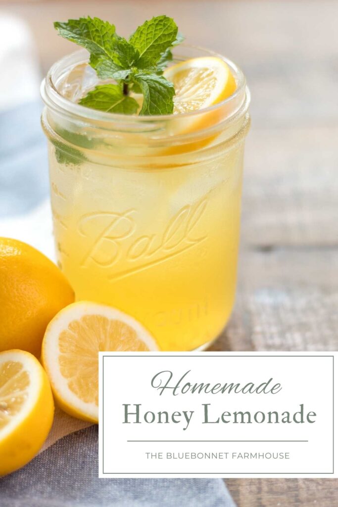 closeup of a glass wide mouth mason jar with iced lemonade garnished with a fresh mint sprig and lemon slice. A whole and halved lemon sitting on a blue and white flour sack towel next to the cup of lemonade. text reads "homemade honey lemonade - the bluebonnet farmhouse"