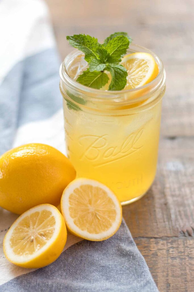 closeup of a glass wide mouth mason jar with iced homemade honey lemonade garnished with a fresh mint sprig and lemon slice. A whole and halved lemon sitting on a blue and white flour sack towel next to the cup of lemonade