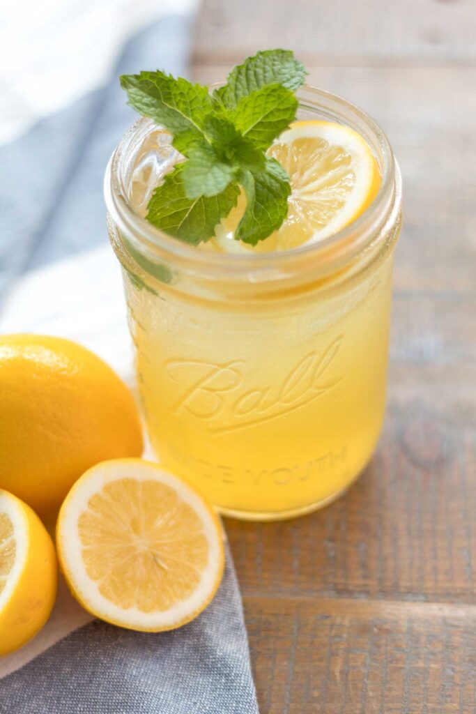 closeup of a glass wide mouth mason jar with iced homemade honey lemonade garnished with a fresh mint sprig and lemon slice. A whole and halved lemon sitting on a blue and white flour sack towel next to the cup of lemonade