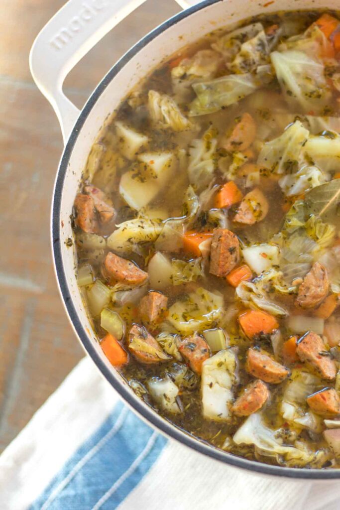 dutch oven full of sausage, cabbage, and potato soup with a white and blue striped tea towel on a farmhouse wood table
