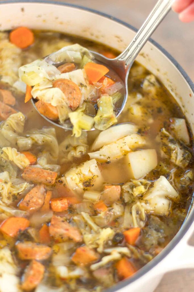 close up of a ladle filled with sausage, cabbage, and potato soup held above a dutch oven