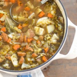 dutch oven full of sausage, cabbage, and potato soup with a vintage ladle and blue striped tea towel on a farmhouse wood table