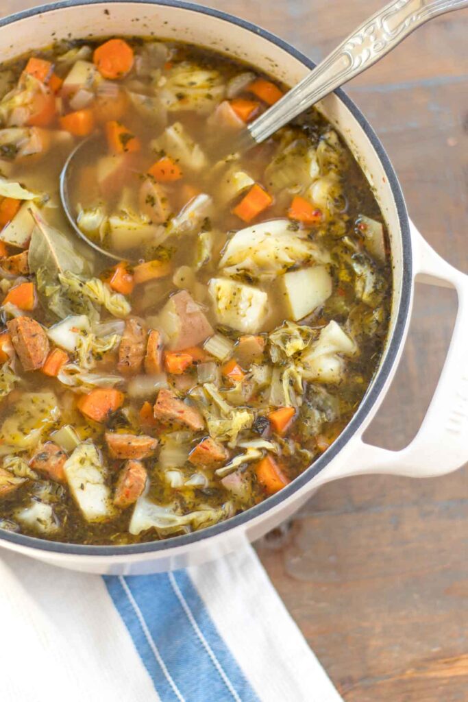 dutch oven full of sausage, cabbage, and potato soup with a vintage ladle and blue striped tea towel on a farmhouse wood table