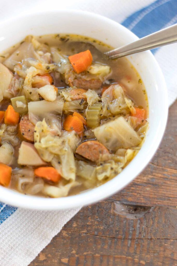 close up of sausage, cabbage, and potato soup in a white bowl with spoon and blue striped tea towel on a farmhouse wood table