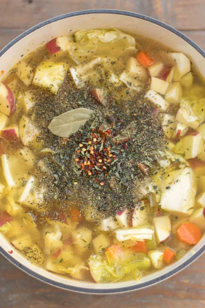 cast iron dutch oven filled with chopped vegetables, sausage, and homemade chicken broth sprinkled with herbs, red pepper flakes, and a bay leaf