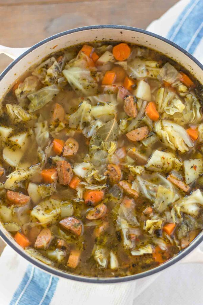 cast iron dutch oven filled with cooked sausage, cabbage, and potato soup consisting of homemade chicken broth, chopped vegetables, herbs, red pepper flakes, and a bay leaf.