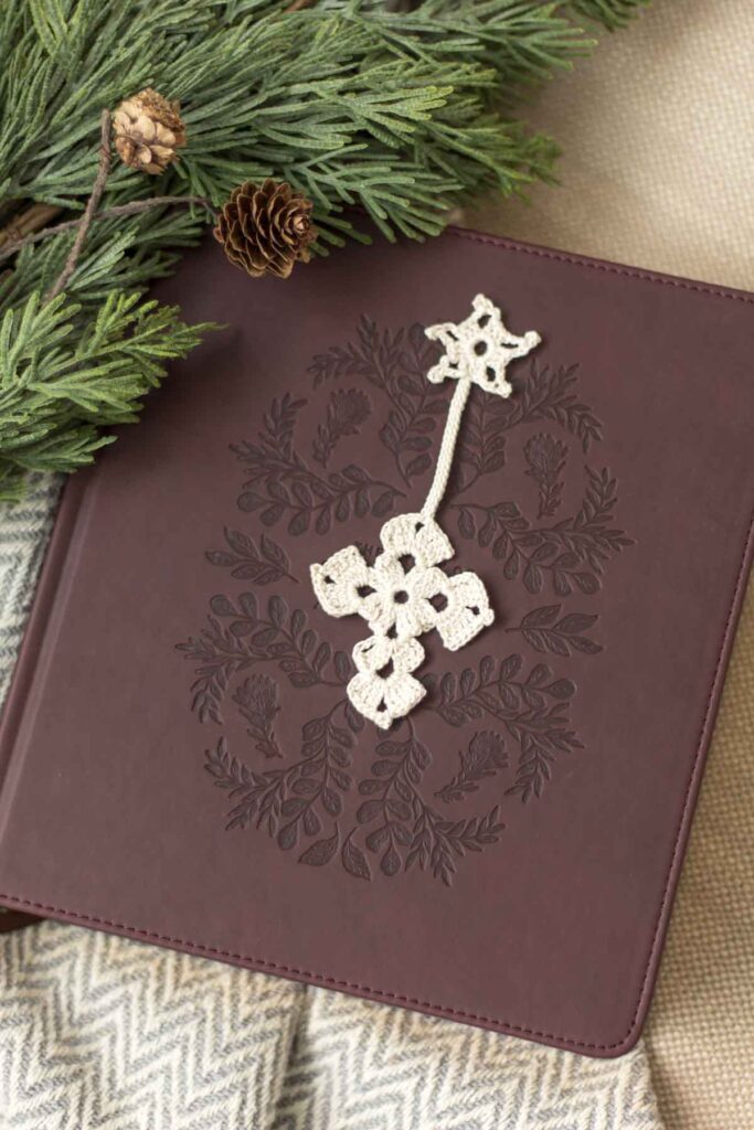 antique white crochet thread Christmas star and cross bookmark on top of a dark brown leather bible with cedar greenery and pine cones next to it on a beige couch with woven neutral blanket