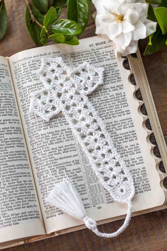 flat lay of a white lace crochet thread cross bookmark with tassel lying on top of an open bible in Mark chapter 15 next to a gardenia on top of a reclaimed farmhouse table